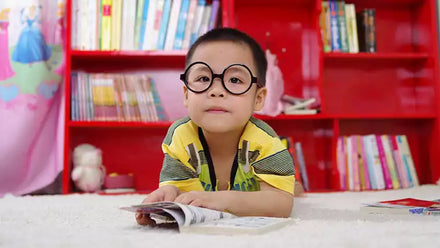 young boy reading book near red shelves